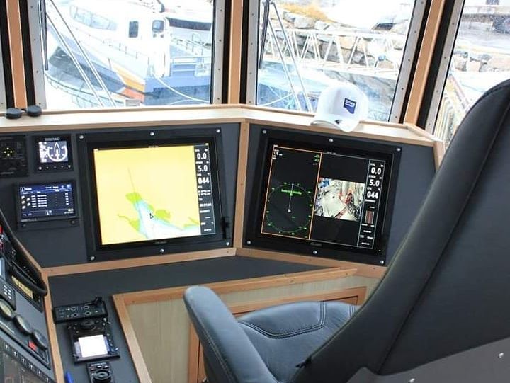 Bilde av en skipsbro med kontrollpanel, to store skjermer som viser navigasjonsdata, og en grå stol foran. Utsikt mot havnen og andre båter er synlig gjennom vinduene.